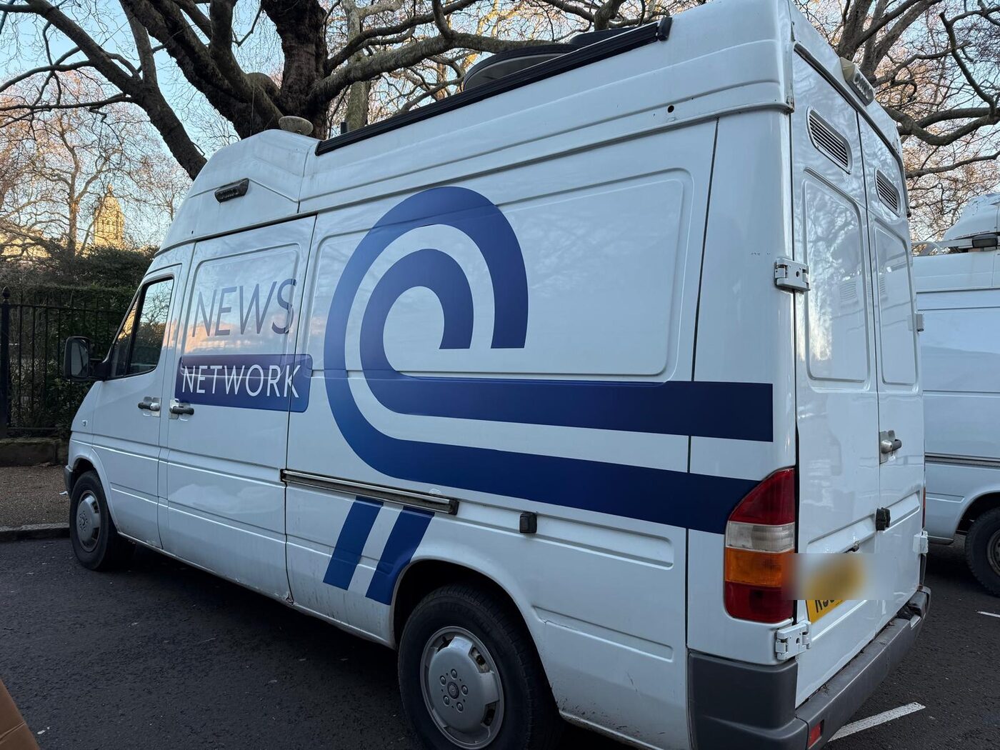 The van in a full fictional News Network wrap with a blue swirl logo, parked under trees in central London