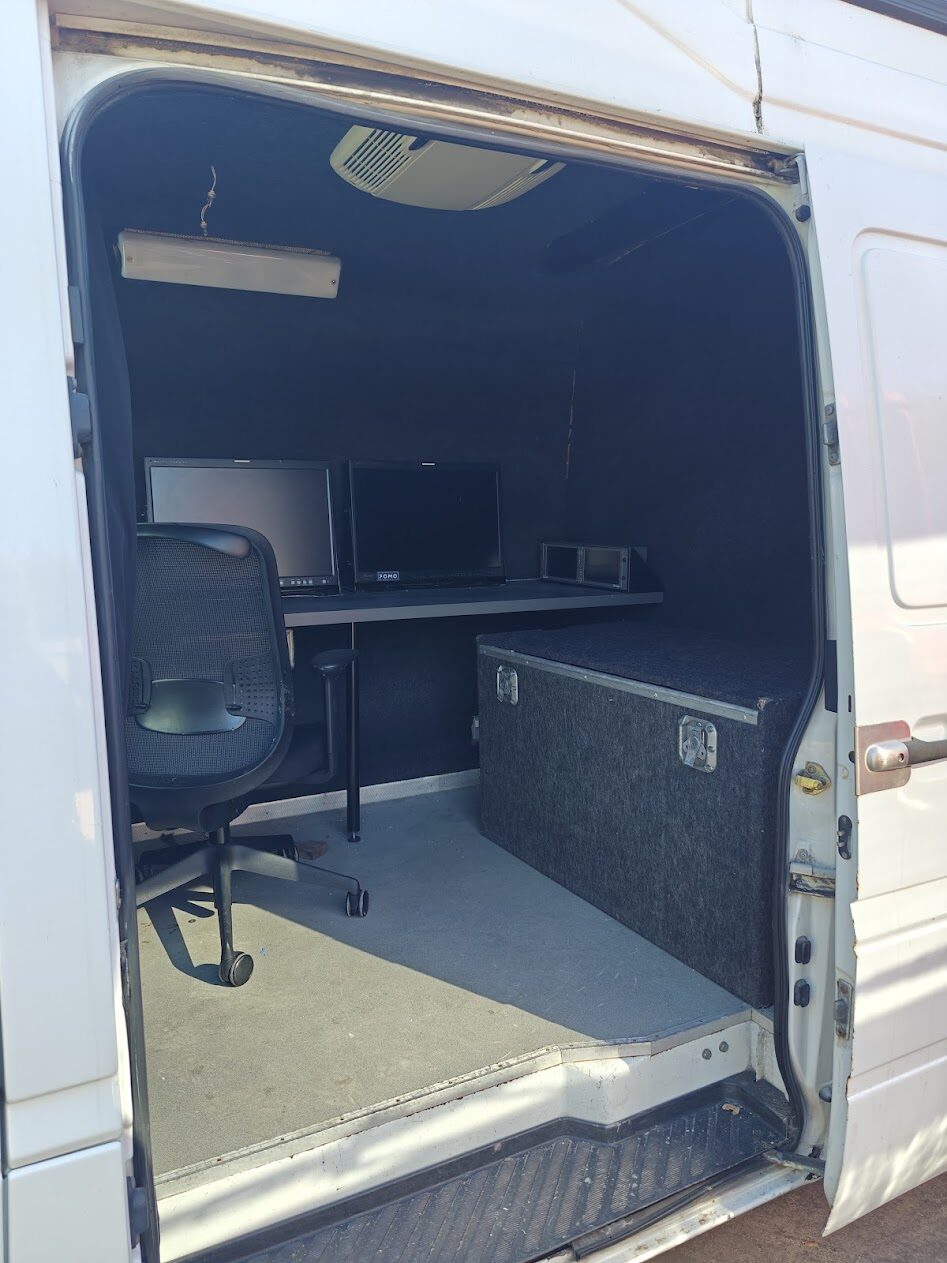 Inside the van's side door: a desk with two monitors, rack unit, operator chair and a large equipment trunk