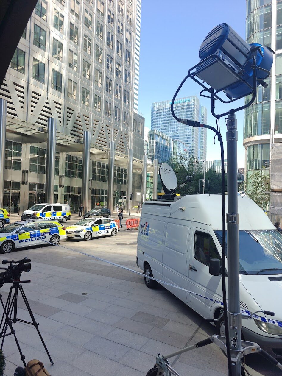 The van with dish raised at Canary Wharf, playing alongside dressed police vehicles with an ARRI lamp rigged in the foreground