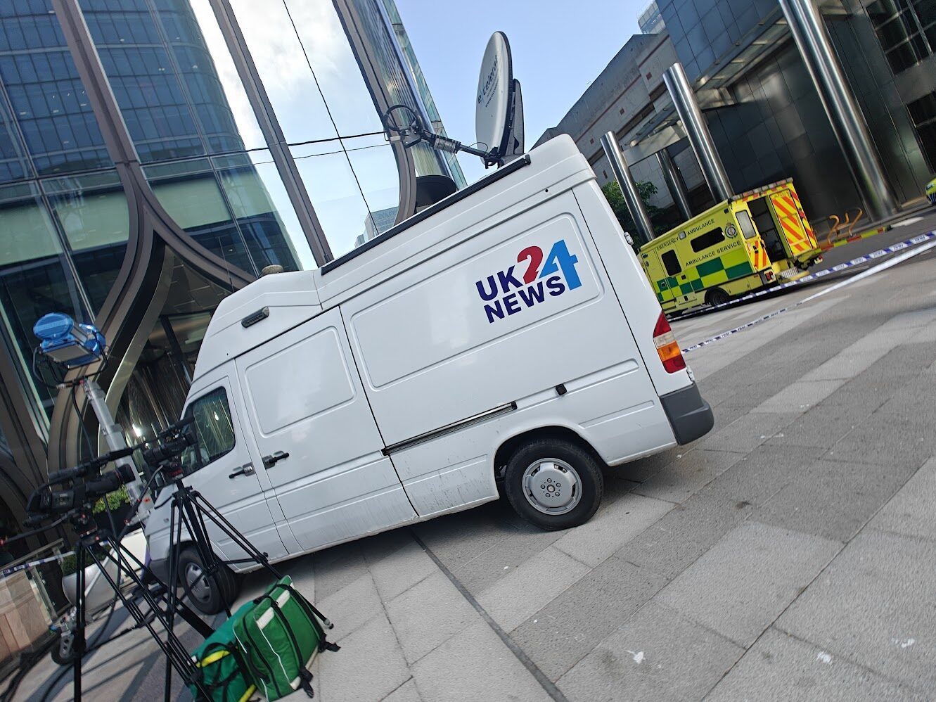 Low dutch-angle shot of the UK24 News van with dish deployed, a dressed ambulance in the background