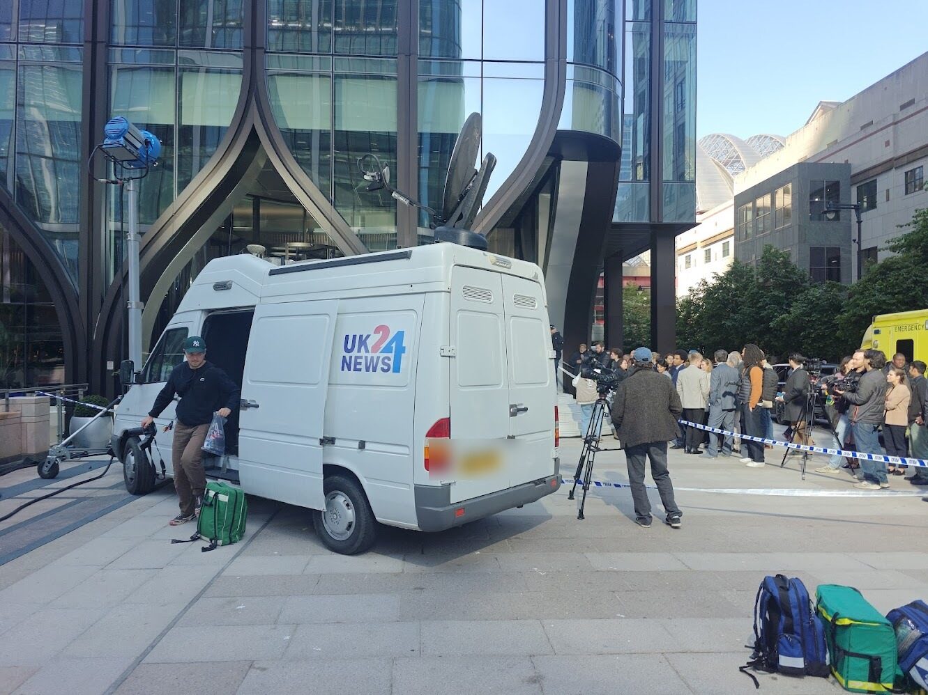 The van dressed as fictional network UK24 News on a drama set at Canary Wharf, crew unloading kit with a press pack gathered behind police tape